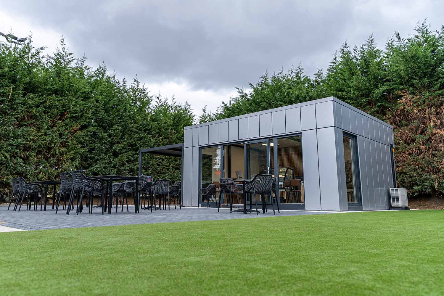 Grey-clad building with glazed doors, an outdoor seating terrace, pergola and an external heat-pump unit.